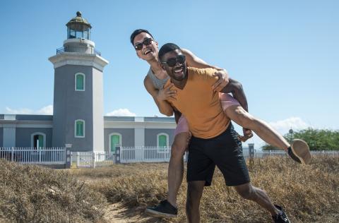 a man giving another man a piggyback ride on a beach in front of a lighthouse