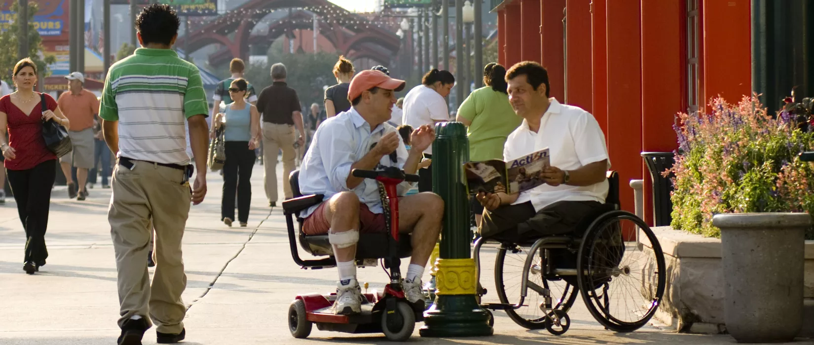 two men in wheelchairs converse in an outdoor setting with many people walking by