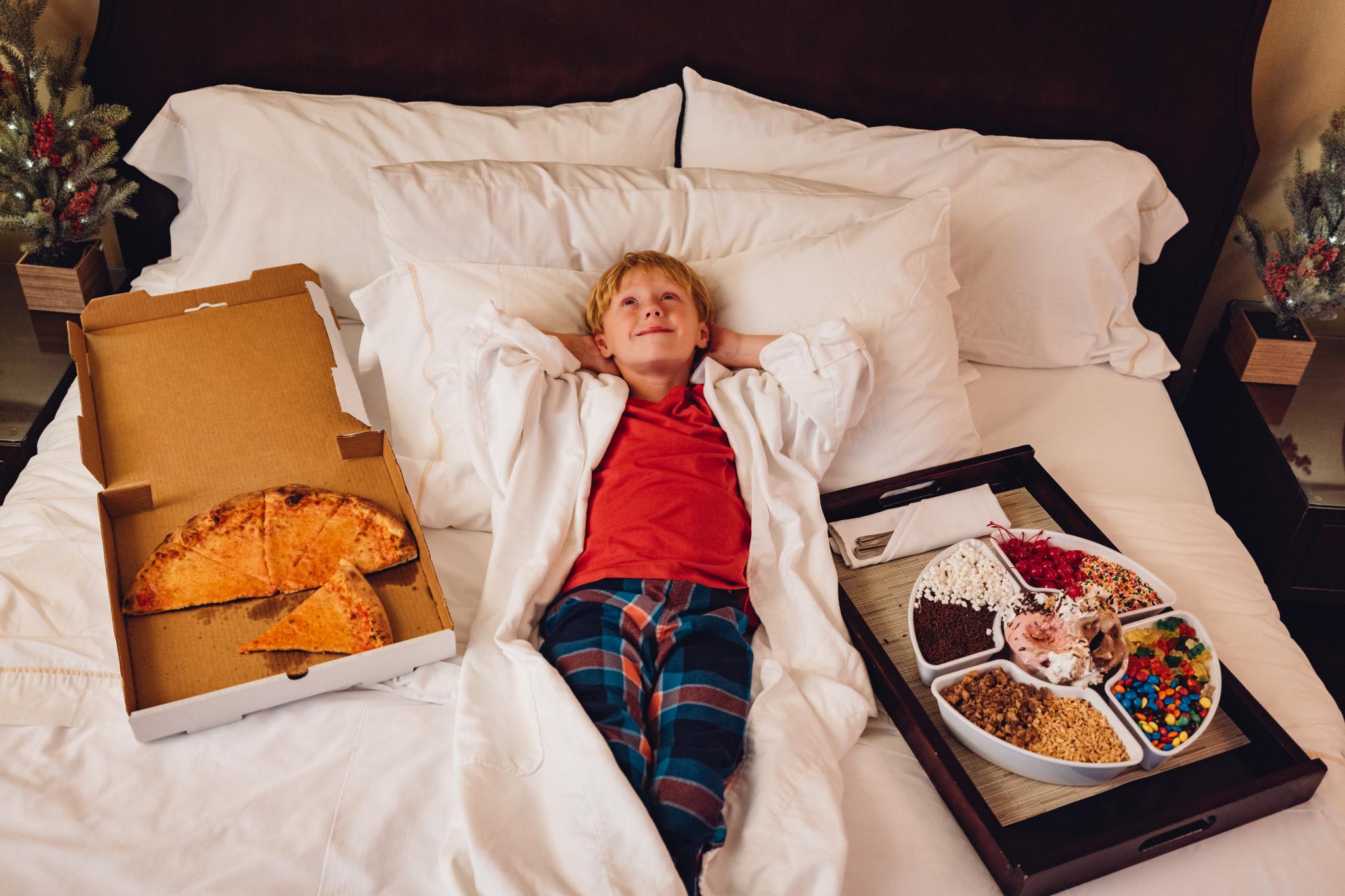 Boy lying in hotel bed surrounded by pizza and snacks