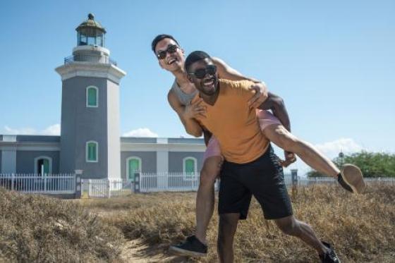 a man giving another man a piggyback ride on a beach in front of a lighthouse