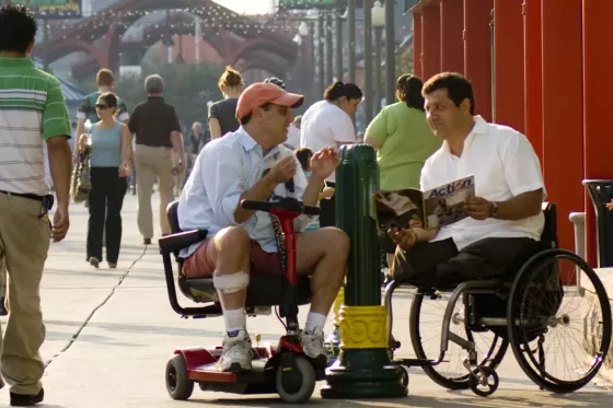 two men in wheelchairs converse in an outdoor setting with many people walking by