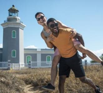 a man giving another man a piggyback ride on a beach in front of a lighthouse