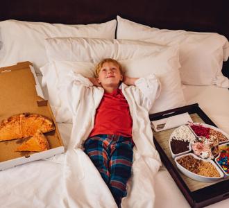 Boy lying in hotel bed surrounded by pizza and snacks